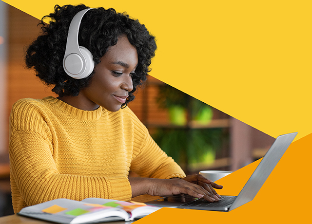 Person wearing headphones and a yellow sweater using a laptop at a desk with an open notebook.